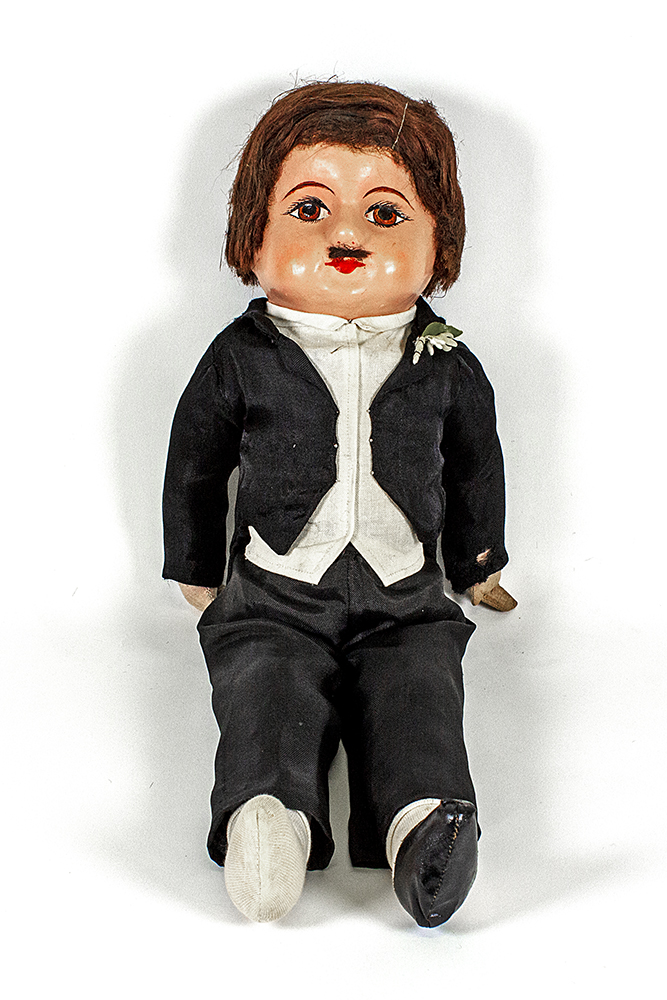 Doll with short brown hair and moustache wearing black suit with fake flower in buttonhole
